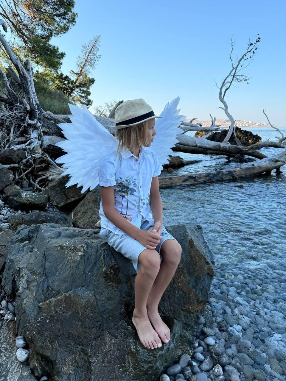 May include: A young person wearing a white shirt and a straw hat sits on a rock by the water. They are wearing large white angel wings.