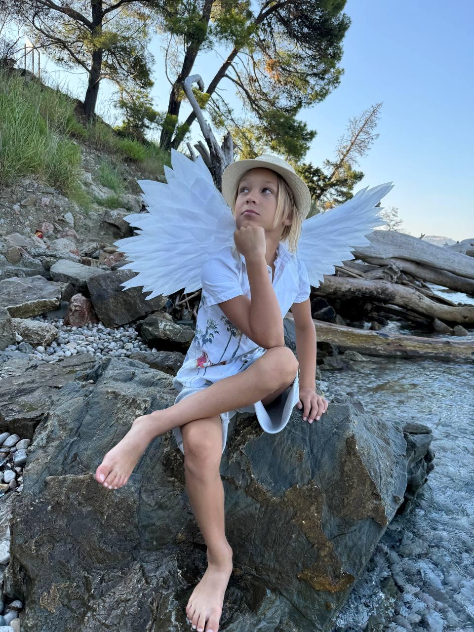 May include: A young person wearing a white shirt and a straw hat with white angel wings sitting on a large rock by a body of water.