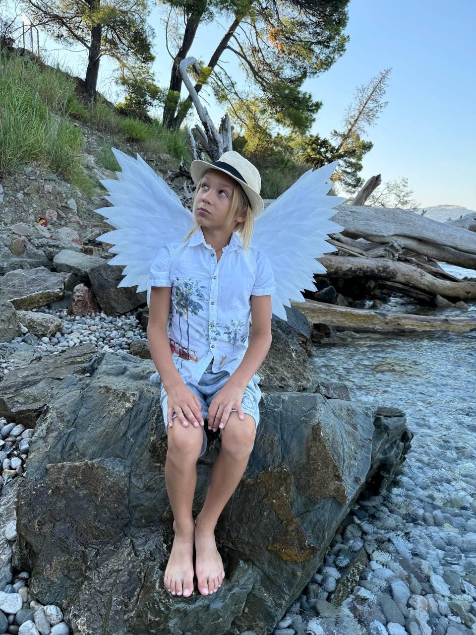 May include: A young person wearing a white shirt and a straw hat sits on a rock by the water. They are wearing white angel wings.