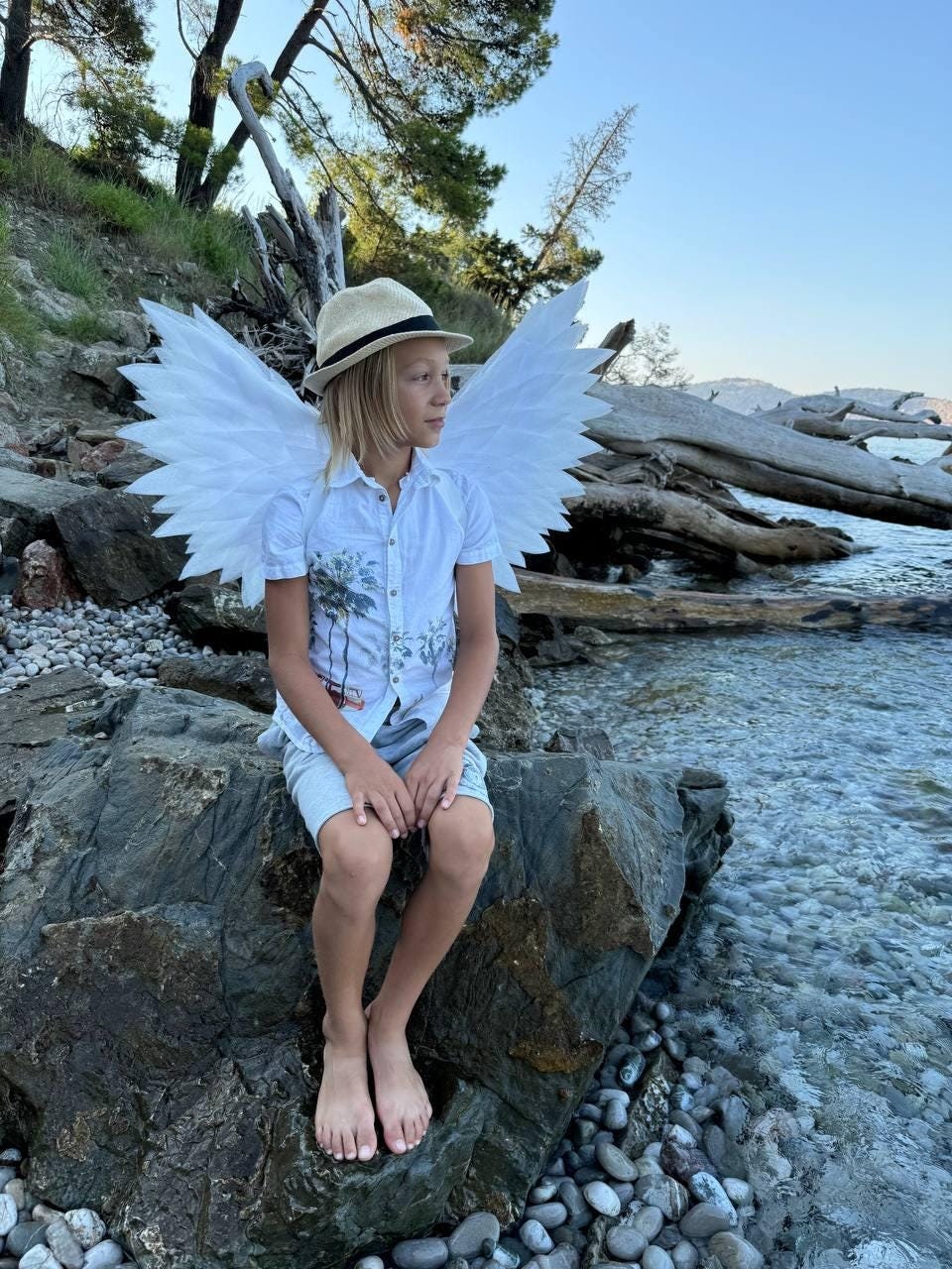 May include: A young person wearing a white shirt and a straw hat sits on a rock by the water. They are wearing large white angel wings.
