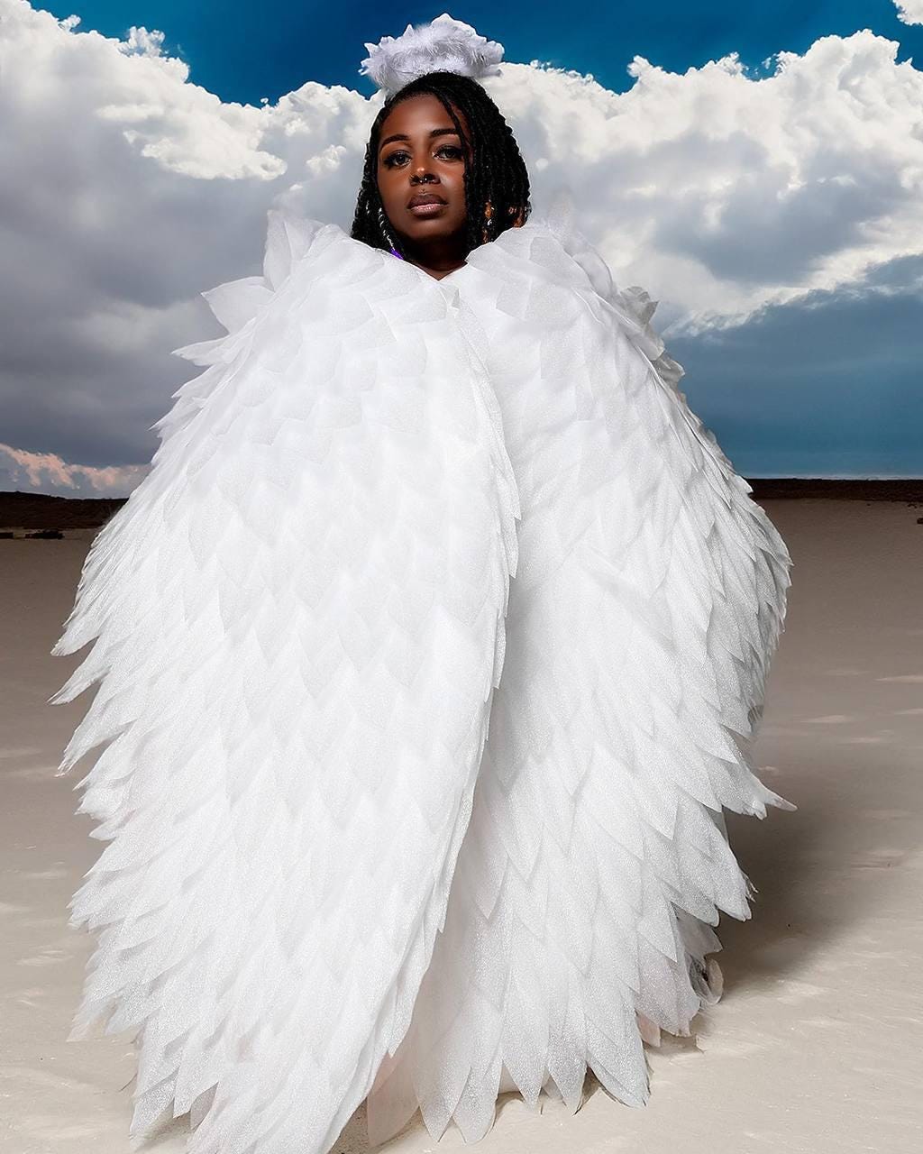 May include: A person wearing large white feathered angel wings and a halo. The wings are the focal point, with a layered design. The background features a cloudy sky and a desert landscape.