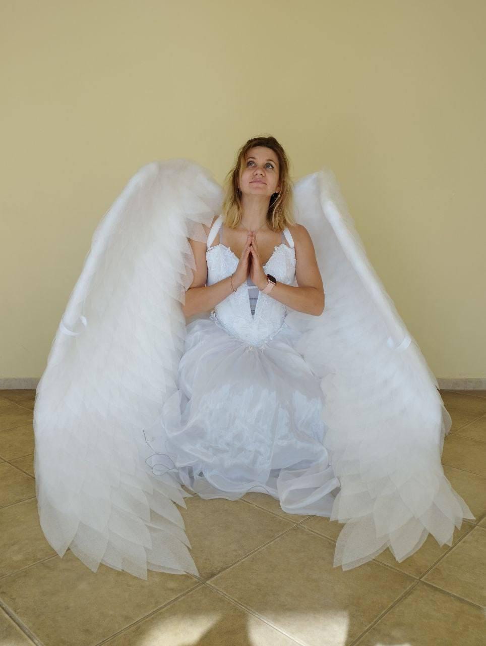 May include: A person wearing a white dress and large white angel wings, kneeling with hands clasped in prayer. The wings are made of layered, feathery material. The dress has a bodice with delicate detailing and a flowing skirt.