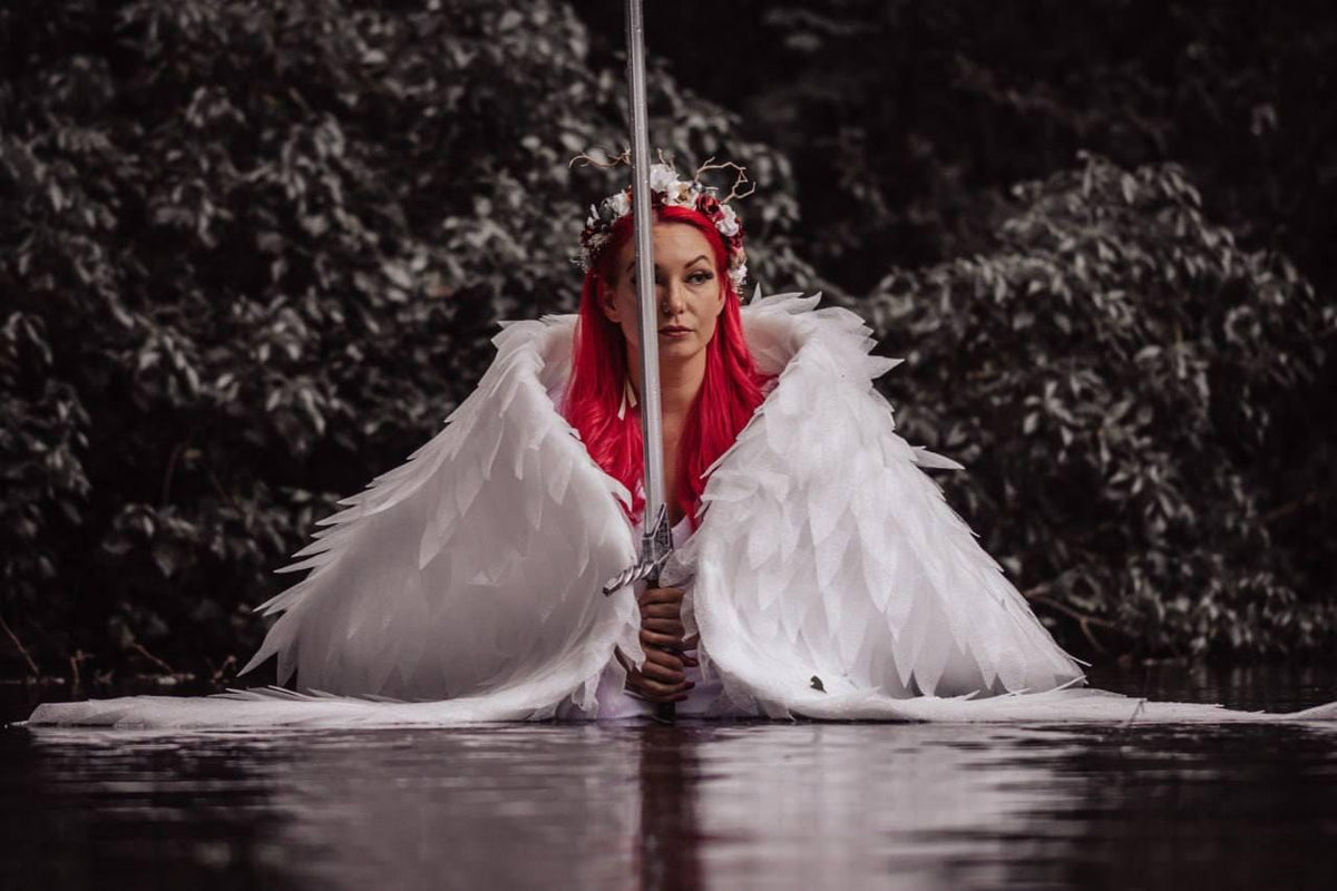 May include: A person with red hair, wearing white feathered angel wings and a floral crown, holds a silver sword. The person is standing in water, with a dark, blurred background of foliage.