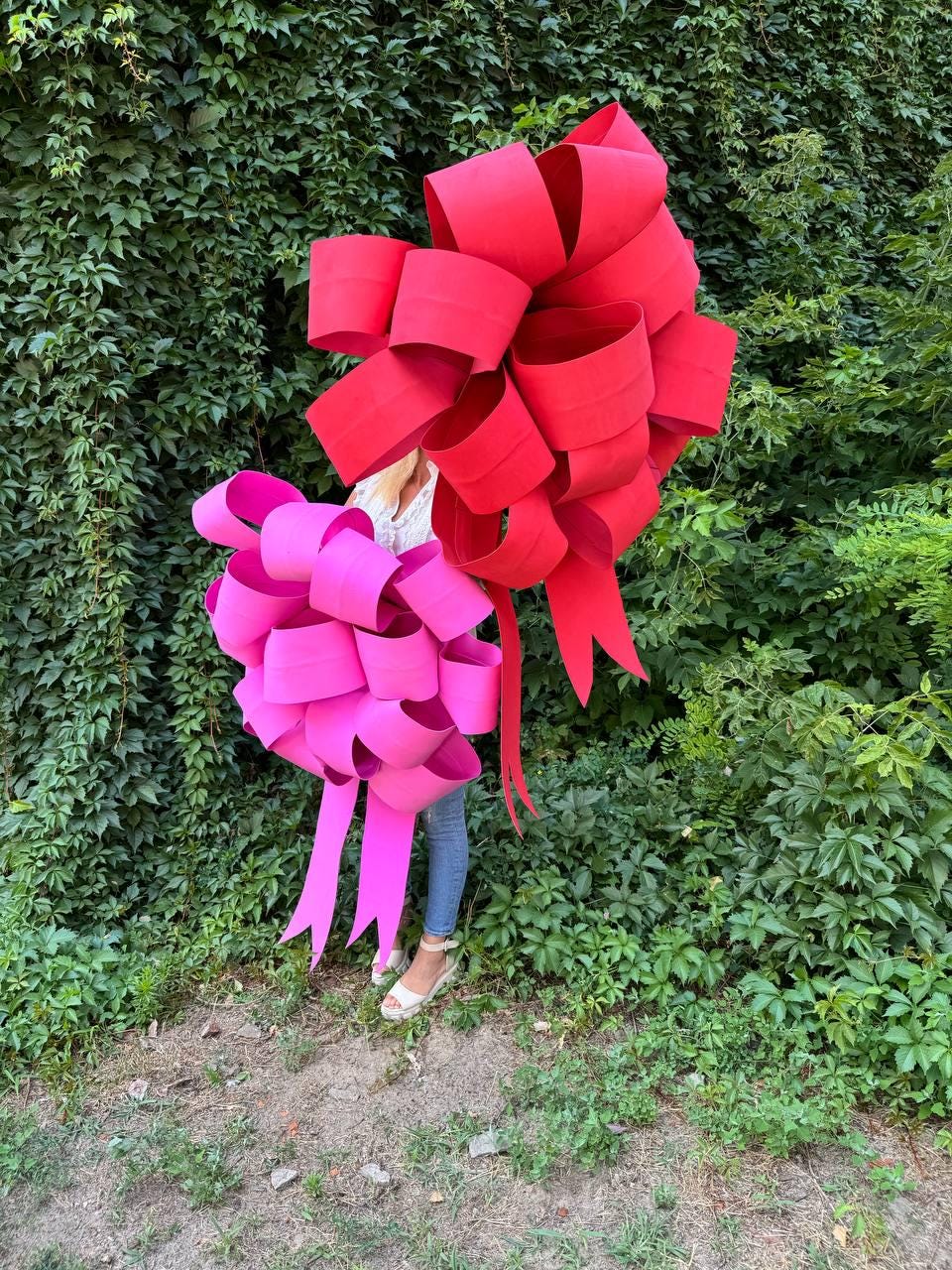 May include: Two large decorative bows, one vibrant red and the other bright pink, are displayed against a backdrop of lush green foliage. The bows have multiple loops and long, flowing tails, suggesting a festive or celebratory theme.