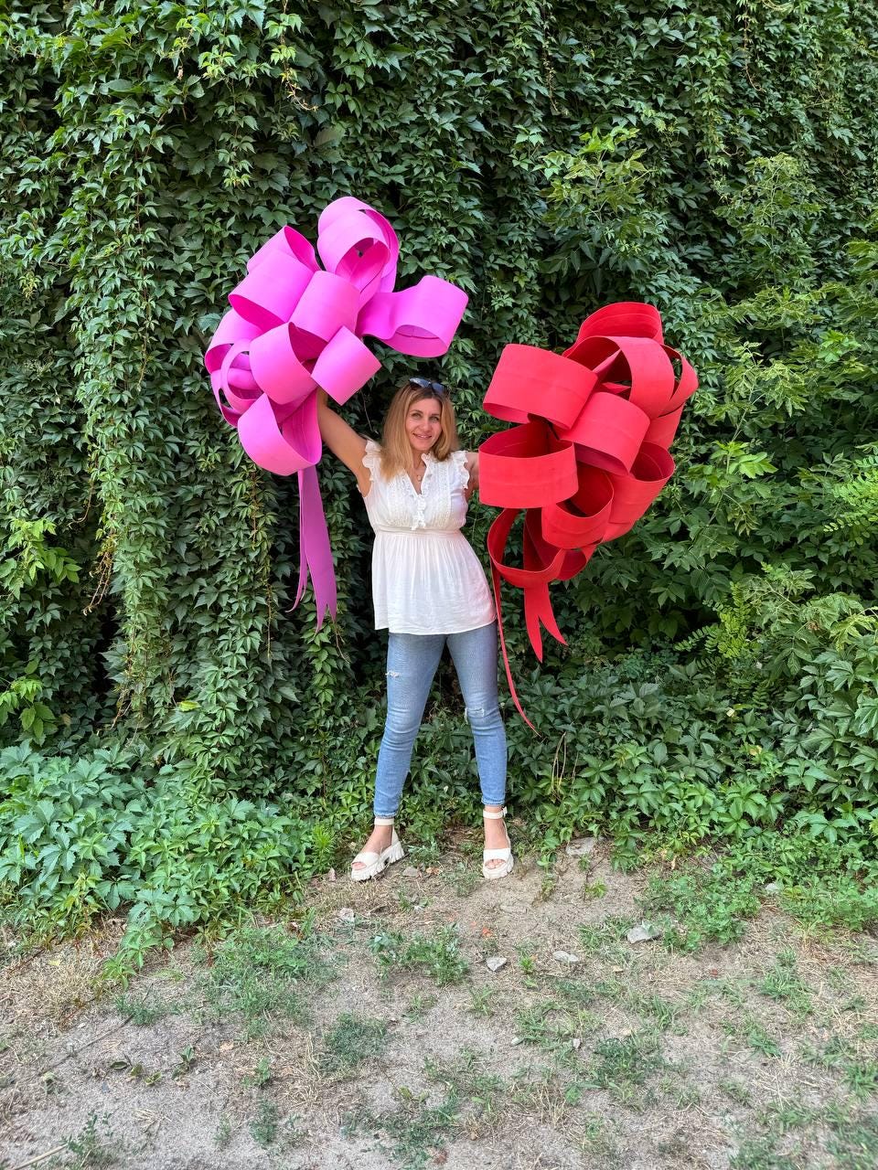 May include: A woman holding two large paper bows. One bow is bright pink, and the other is red. The bows are made of folded paper strips and have long, flowing ribbons. The background is a wall of green foliage.