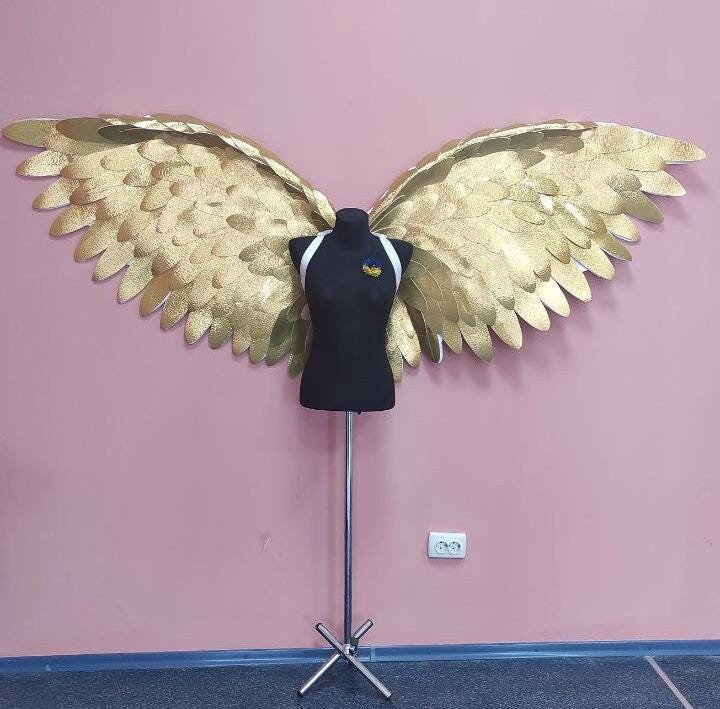May include: Large, golden angel wings with a textured surface are displayed behind a black mannequin. The mannequin has white straps and a small pin with blue and yellow colors. The wings are mounted on a metal stand against a pink wall.