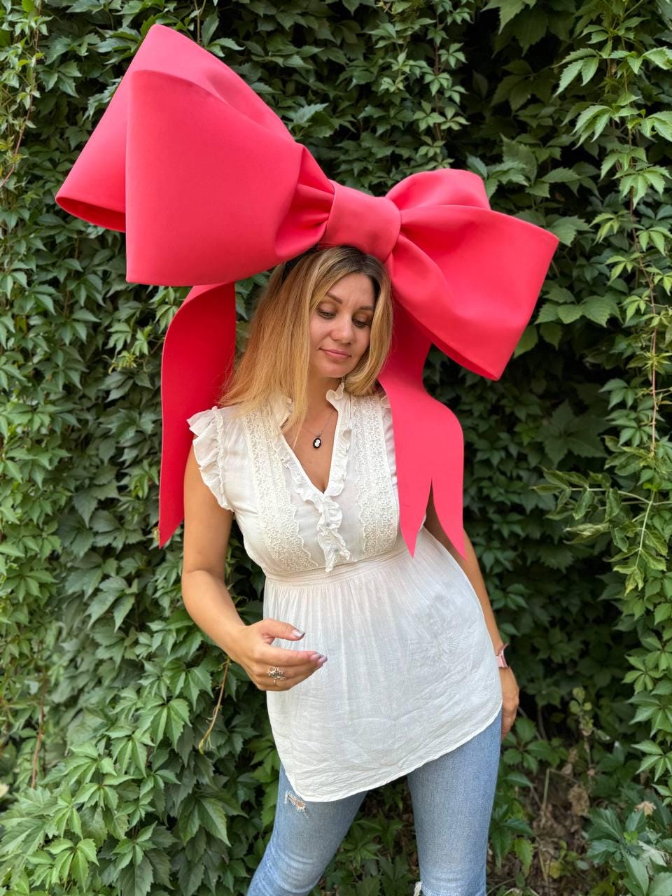 May include: A woman wearing a white top and blue jeans is standing in front of a green leafy background. She is wearing a large, bright pink bow on her head.