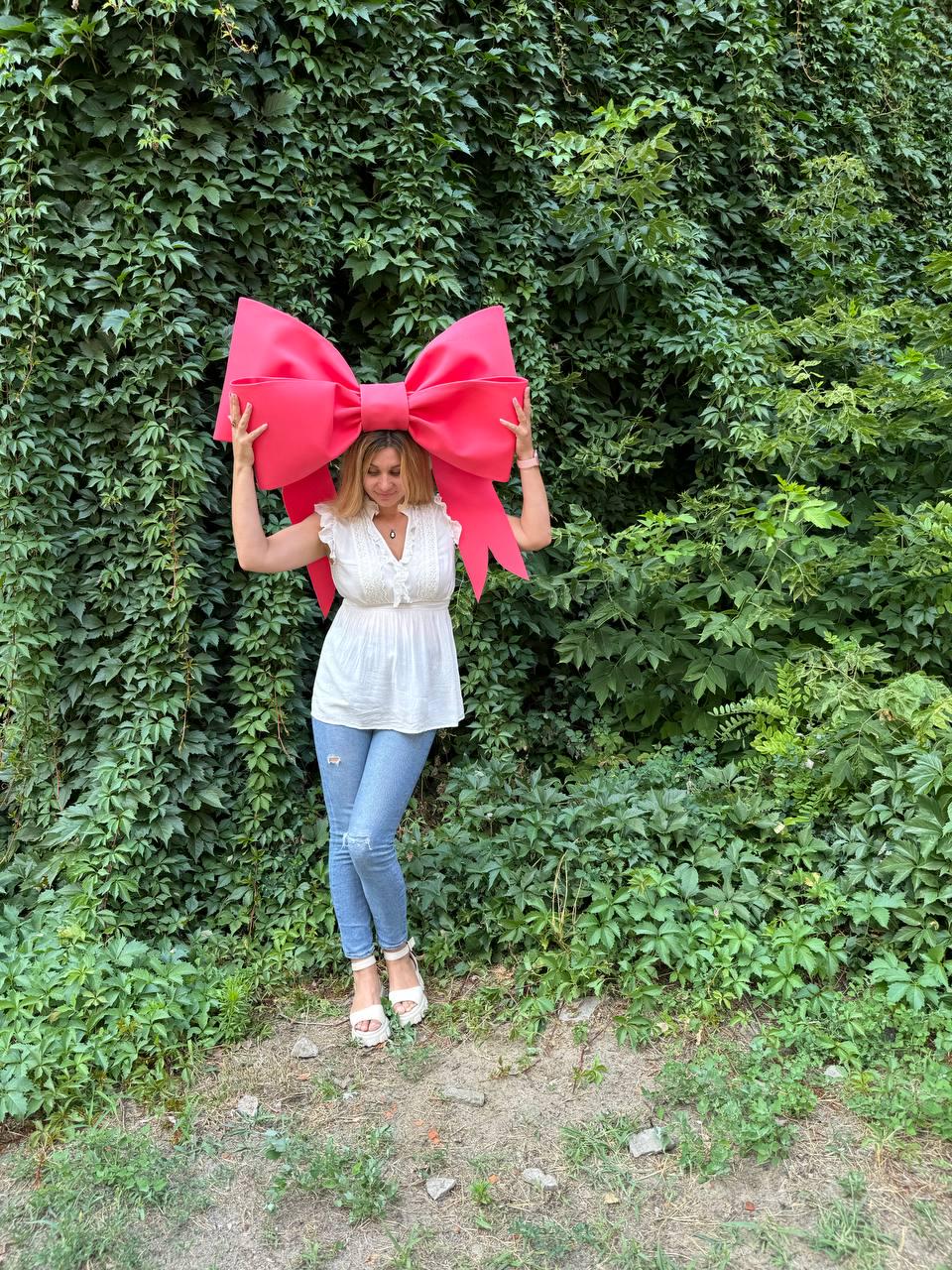 May include: A woman is holding a large, bright pink bow over her head. She is wearing a white blouse, blue jeans, and white sandals. The bow is in front of a green leafy wall.