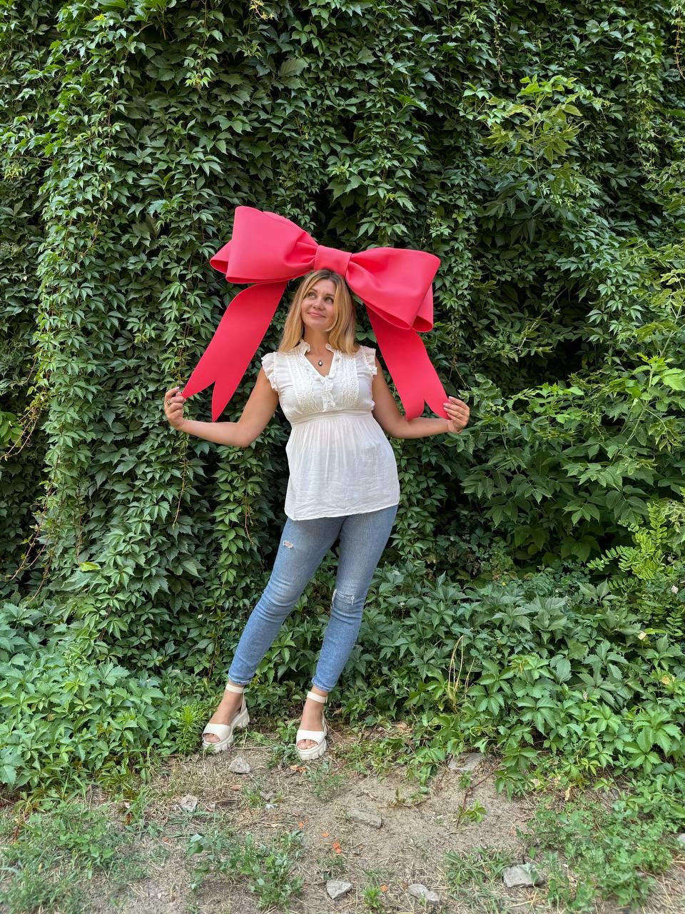 May include: A woman wearing a white top and blue jeans is holding a large, bright pink bow in front of a green leafy wall.
