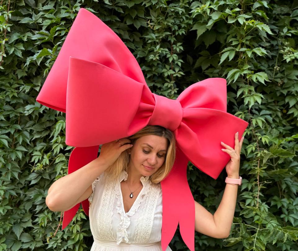 May include: A large, bright pink bow sits on top of a person's head. The bow is made of a soft, fabric-like material and has a large, rounded center.