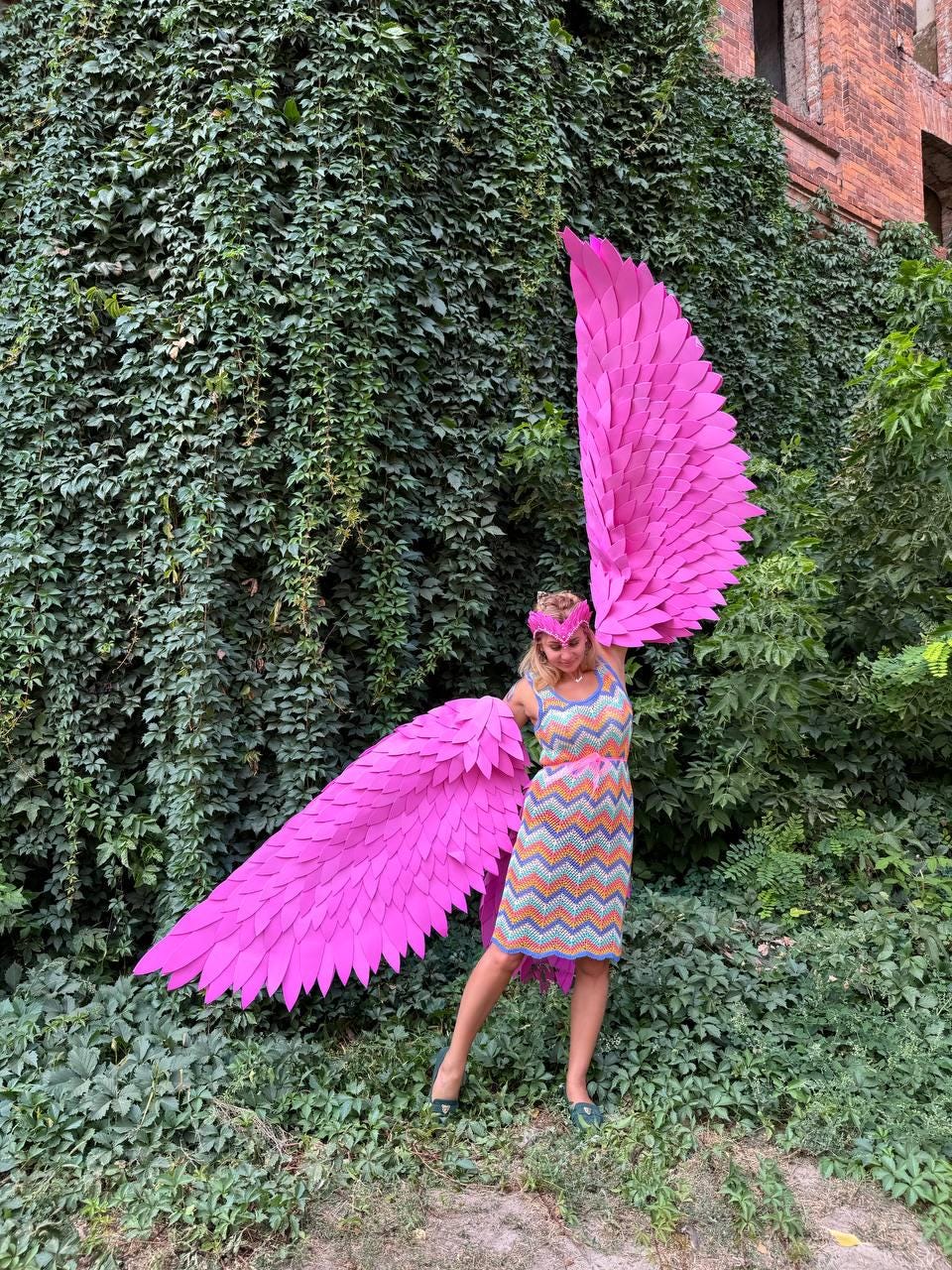 May include: A person wearing a colorful dress and a pink headband holds large pink paper wings in front of a green ivy wall.