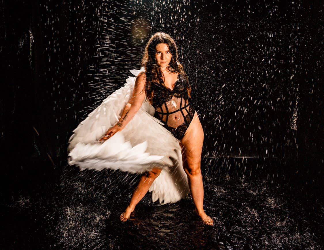 May include: A woman wearing a black lingerie bodysuit with a sheer mesh panel and white angel wings, standing in a dark room with water droplets falling around her.