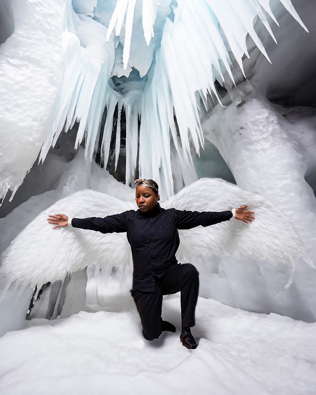 May include: A person wearing a black shirt and pants kneels in a snowy cave with large white wings outstretched. The cave is decorated with icicles hanging from the ceiling.
