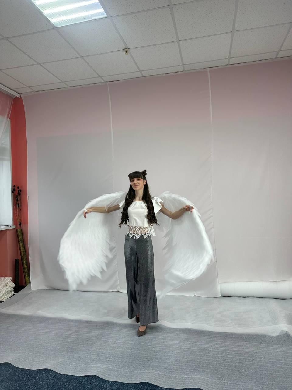 May include: A woman wearing a white top and gray pants with white angel wings. The wings are large and feathery.