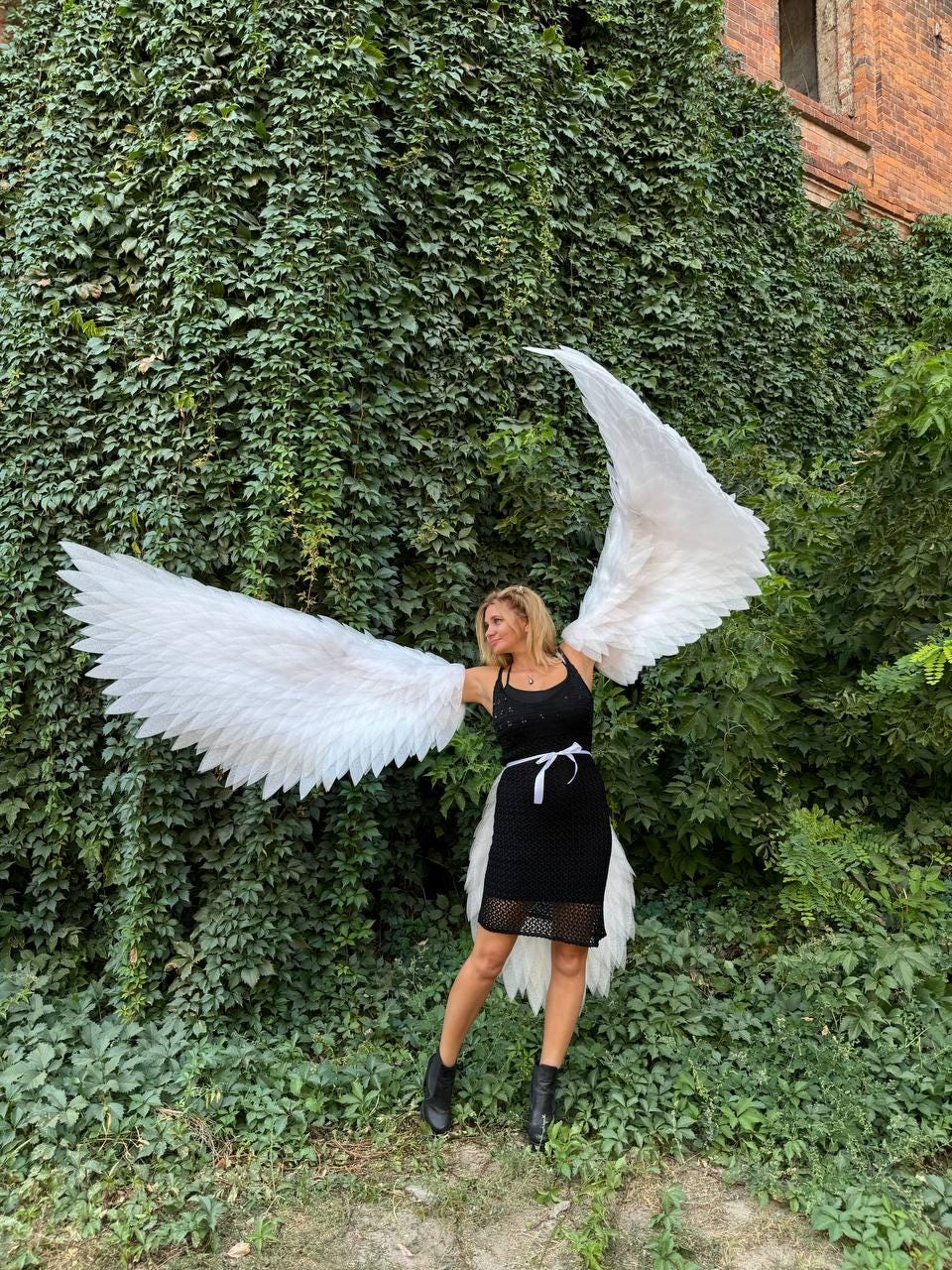 May include: A woman wearing a black dress and white angel wings stands in front of a green ivy wall.