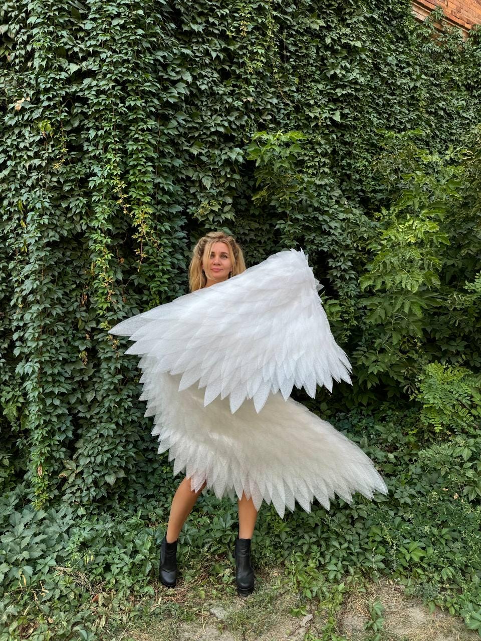 May include: White angel wings with layered, feather-like details. The wings are large and spread out, with a person wearing black boots and standing in front of a wall of green foliage. The wings are the focal point of the image.