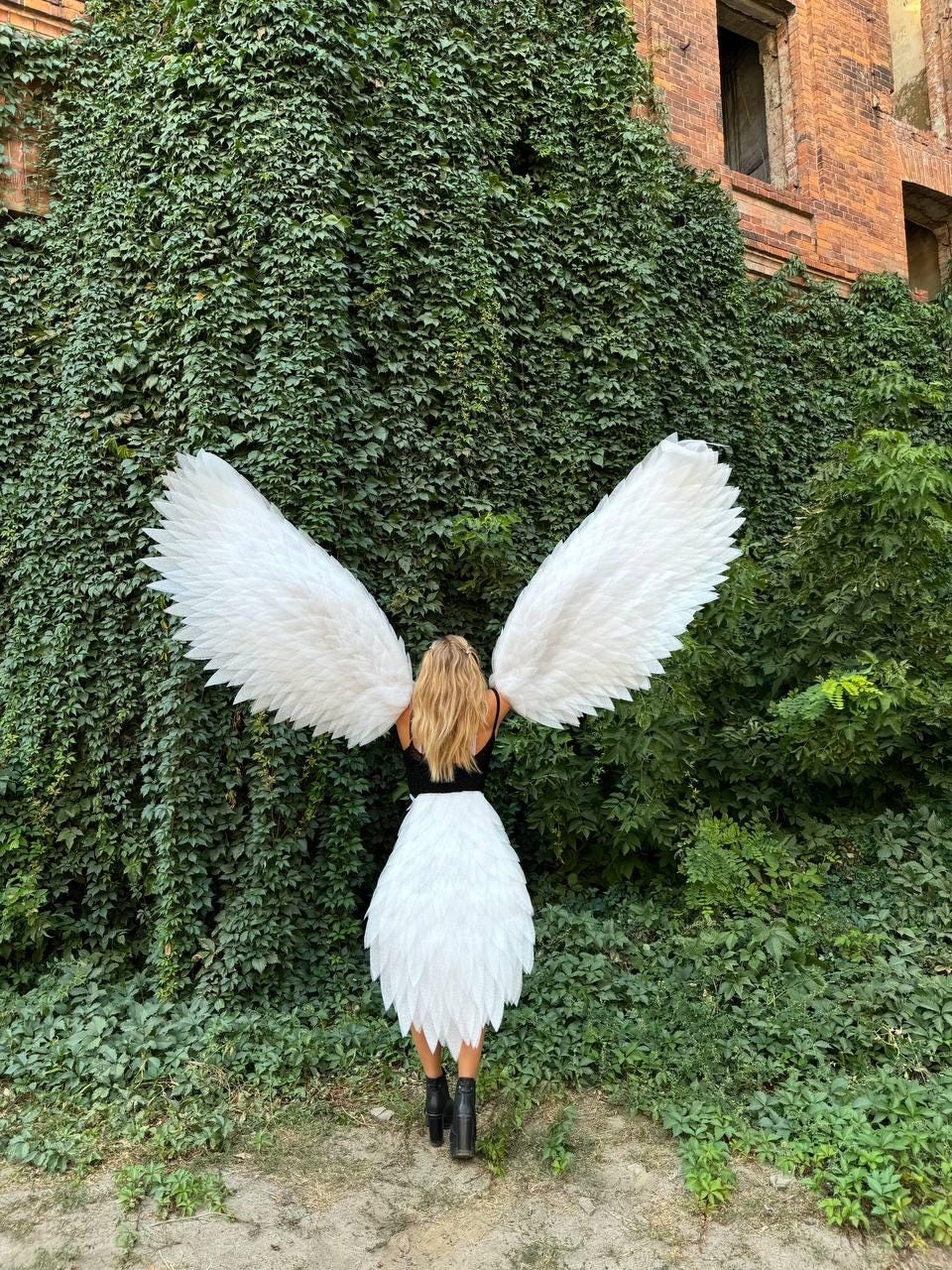 May include: A person wearing a black top and white feathered skirt and large white feathered wings, standing in front of a wall of green ivy. The person is wearing black boots. The wings are spread wide.