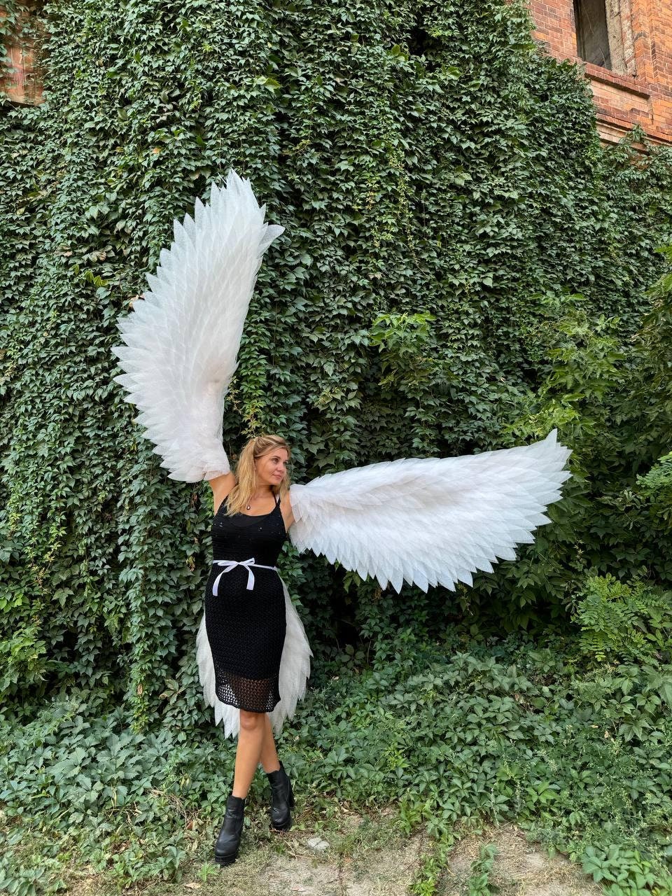 May include: A person wearing a black dress and white angel wings, standing in front of a green ivy covered wall.