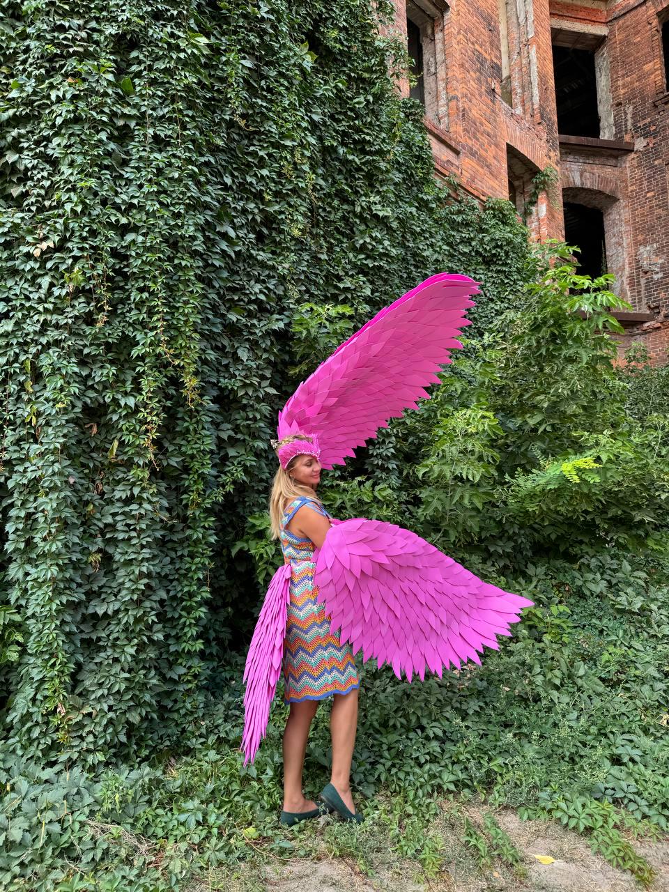 May include: A person wearing a pink feathered angel costume with large wings. The costume is made of pink feathers and is worn over a colorful dress. The person is standing in front of a brick wall covered in ivy.