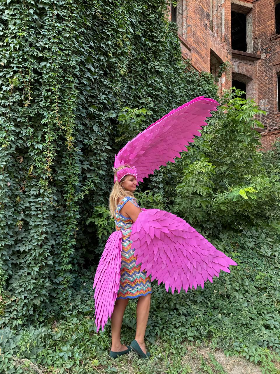 May include: A person wearing a pink costume with large pink wings. The wings are made of feathers and are attached to the person's back. The person is standing in front of a green wall.