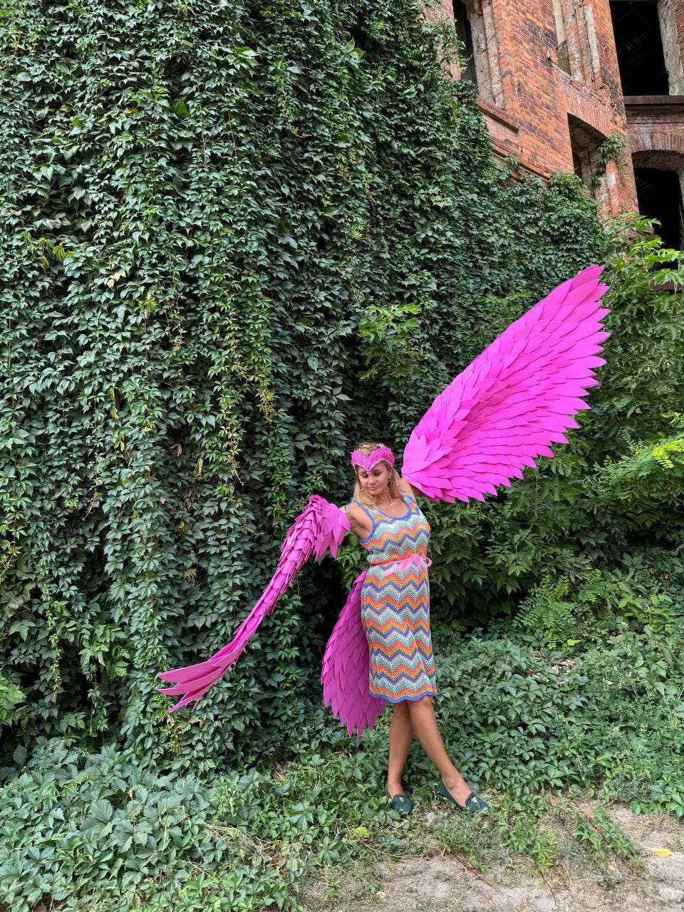 May include: A person wearing a pink and orange patterned dress with large pink wings made of paper. The person is standing in front of a green ivy covered wall.