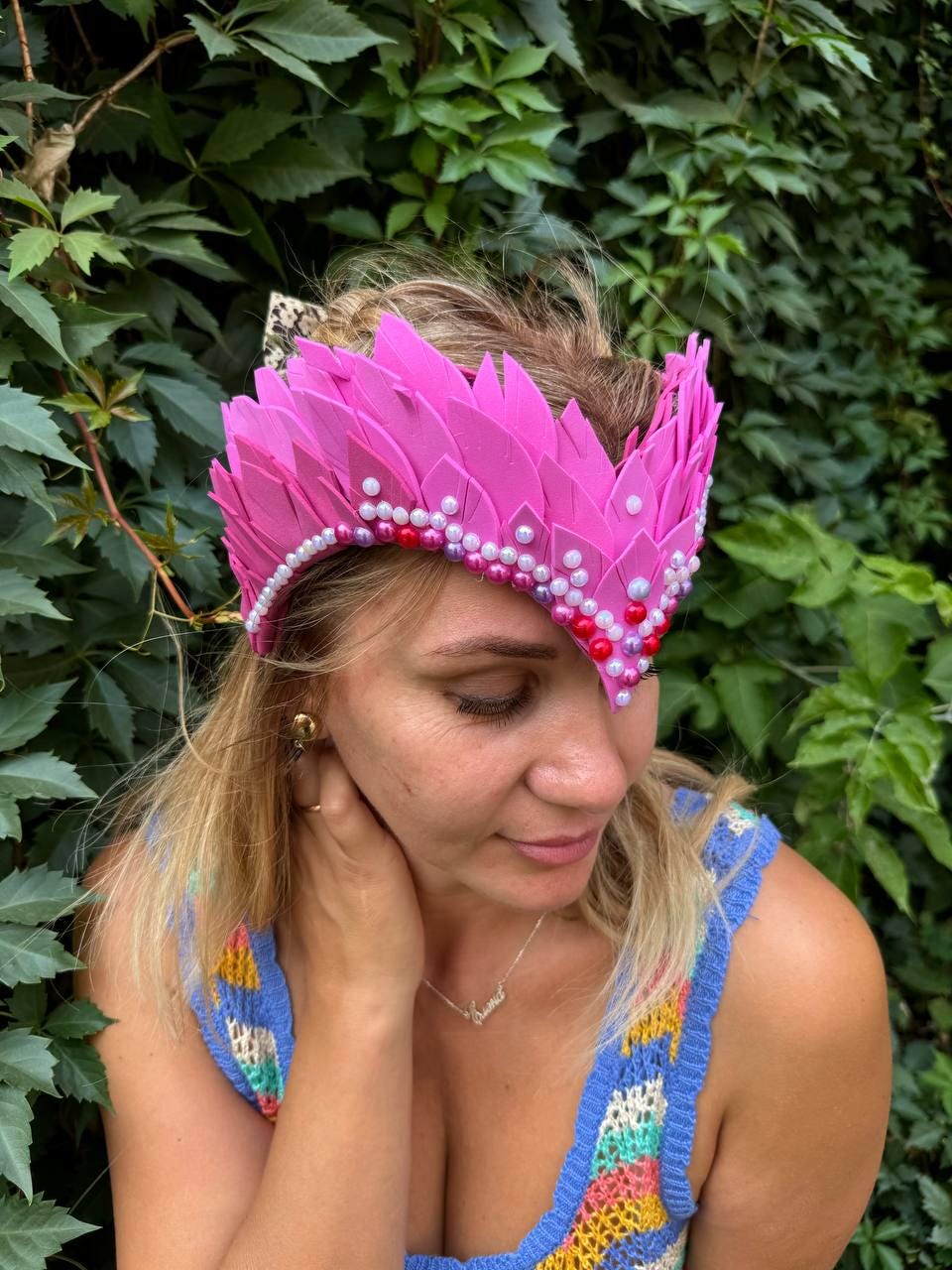May include: A pink feather crown with pearl and red bead embellishments. The crown is worn on a person's head.