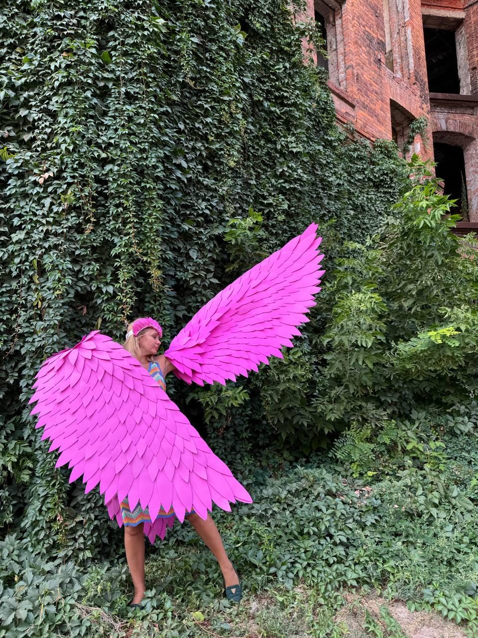 May include: A person wearing a pink costume with large, feathery wings. The wings are made of pink paper and are attached to the person's back. The person is standing in front of a brick wall covered in green ivy.