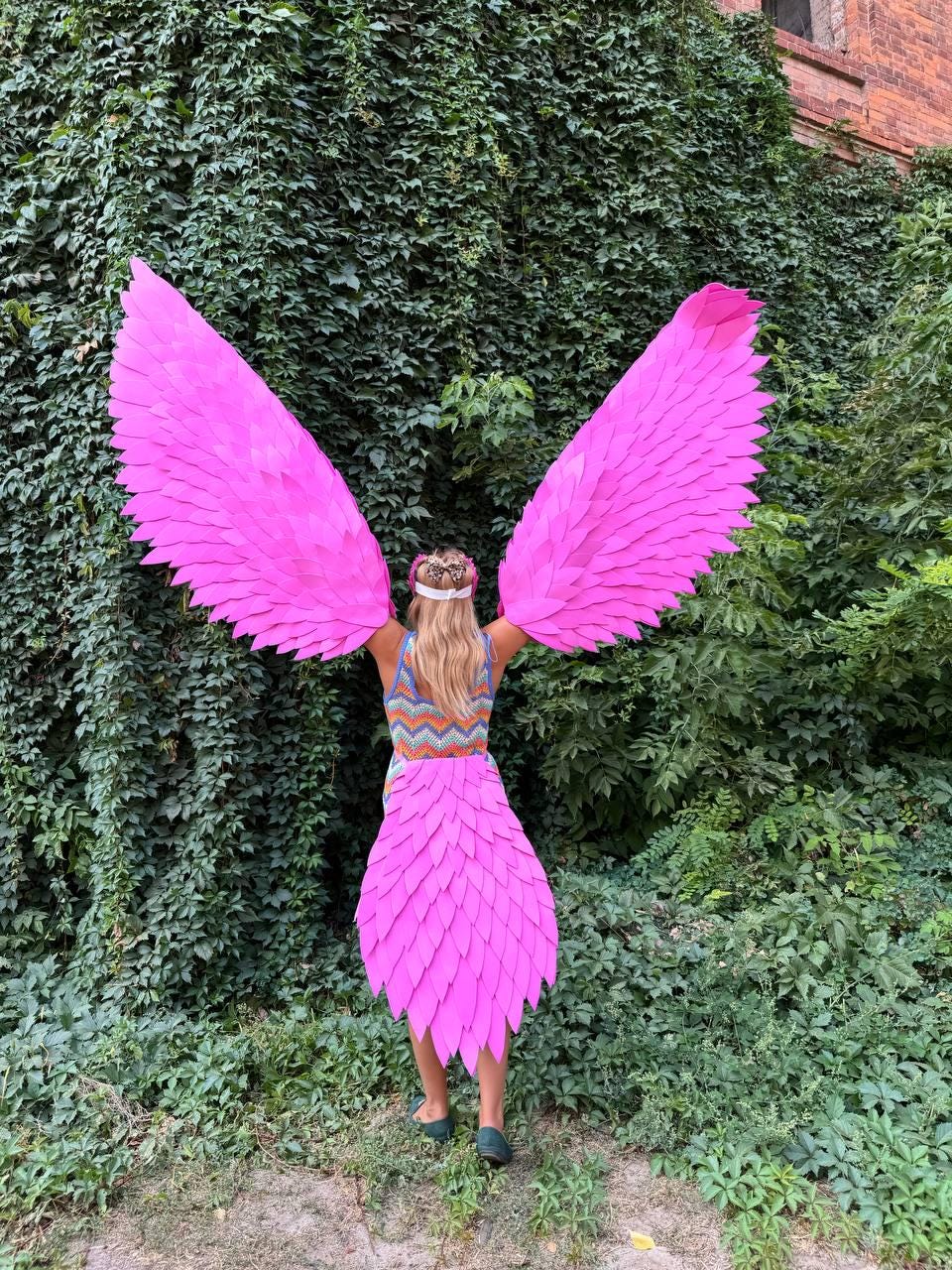 May include: A person wearing a pink feathered angel costume with large wings and a tail. The costume is made of pink feathers and is worn over a colorful patterned shirt.