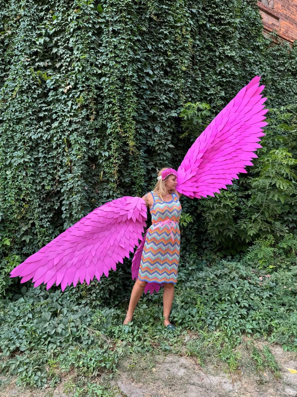 May include: A person wearing a colorful dress and a headband is holding large pink angel wings. The wings are made of feathers and are very detailed.