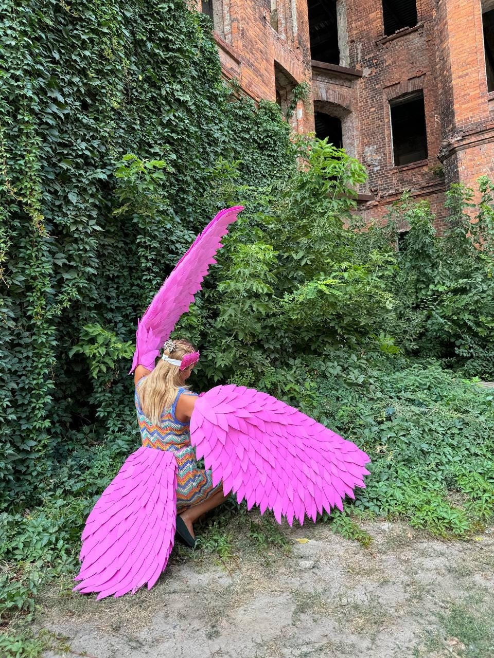 May include: A person wearing a pink angel costume with large, feathery wings. The wings are made of pink fabric and have a layered, textured look. The person is standing in front of a brick wall covered in ivy.