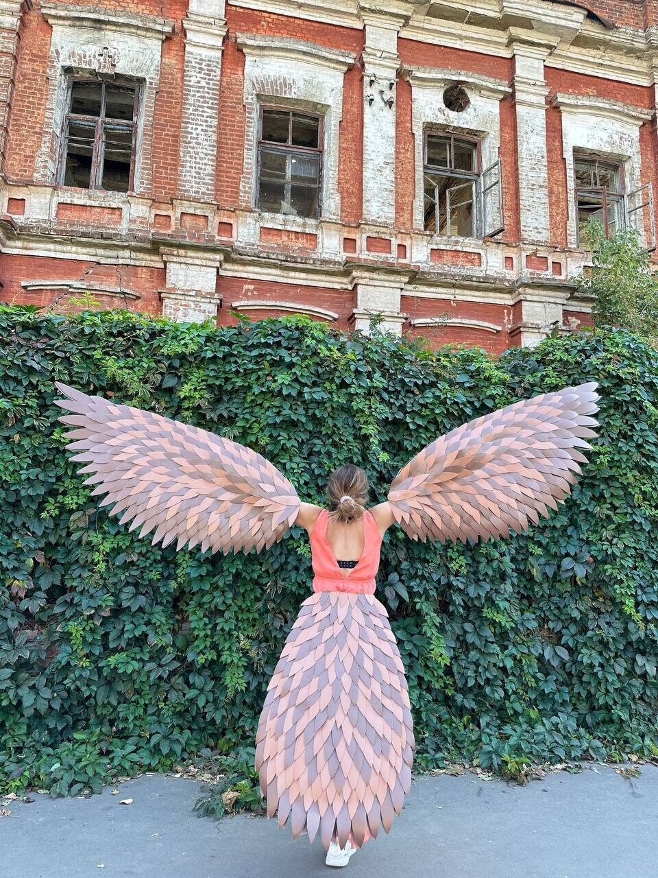 May include: A person wearing a pink and brown feathered costume with large wings, standing in front of a brick building with ivy growing on it.