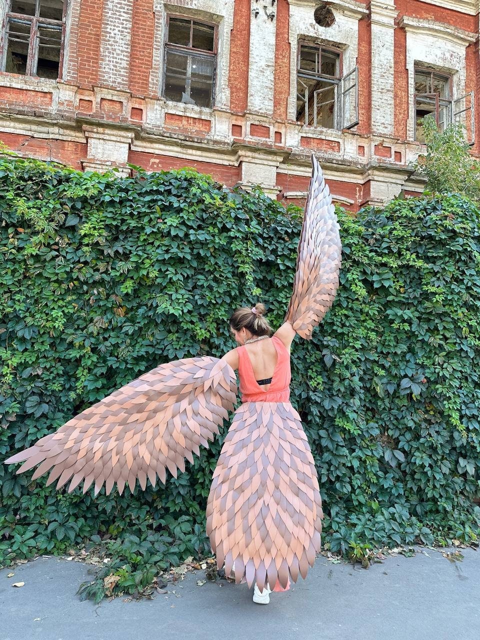 May include: A person wearing a costume with large, brown and tan feathered wings and a matching skirt. The costume is set against a backdrop of green foliage and a weathered brick building.