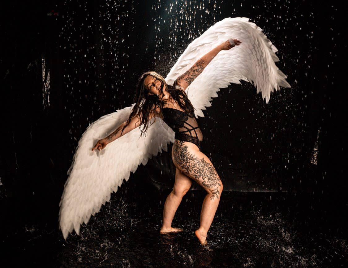 May include: A person wearing a black lingerie outfit with white angel wings, standing in front of a black background with water droplets falling around them.