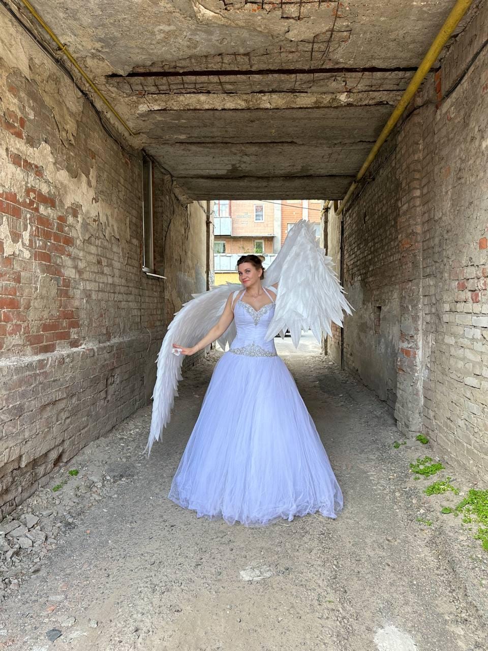 May include: A woman wearing a white dress and large white angel wings stands in a brick alleyway.