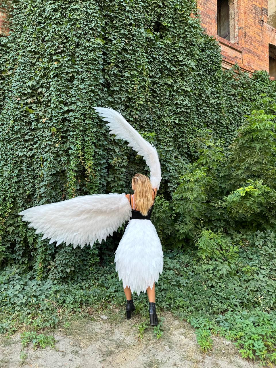 May include: A person wearing a white feathered skirt and large white feathered wings. The person is standing in front of a wall covered in green vines and foliage. The person is wearing a black top and black boots.