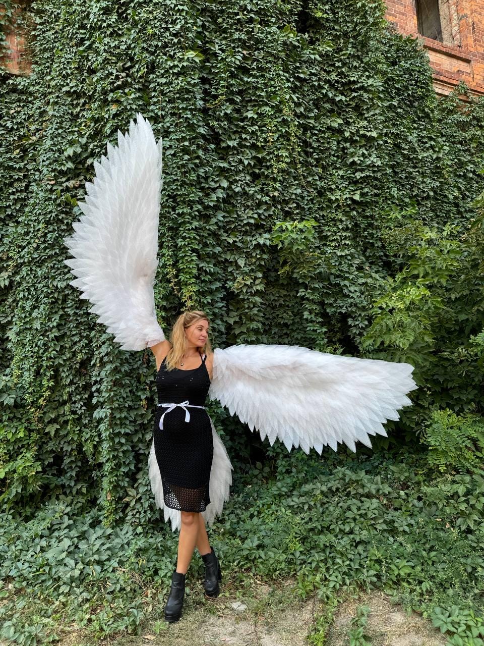May include: A person wearing a black dress and white angel wings, standing in front of a green ivy covered wall.