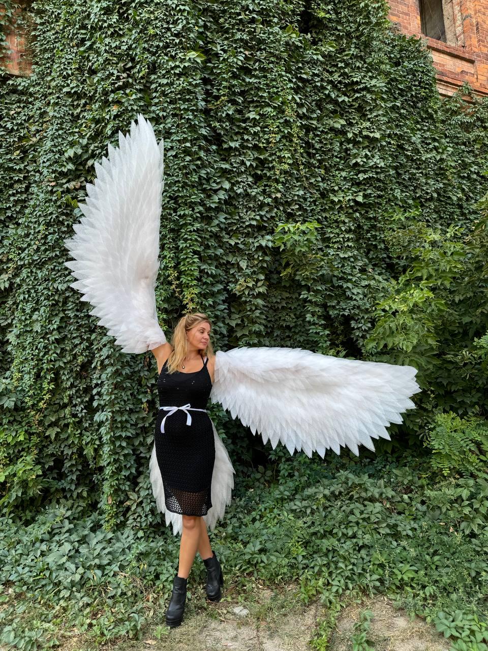 May include: A woman wearing large, white feathered angel wings, standing in front of a wall covered in green ivy. She is wearing a black dress with a white belt and black boots. The wings are spread wide.