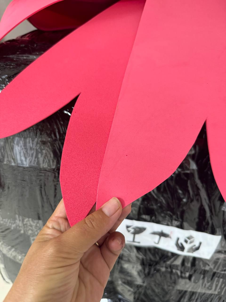 May include: Close-up of a bright pink, petal-shaped foam cutout. The cutout is held by a hand, revealing a glimpse of a black-wrapped object in the background. The foam has a smooth texture and a vibrant color.