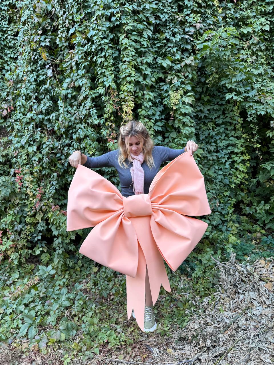 May include: A large peach-colored bow made of fabric. The bow is being held by a person in front of a green leafy background.