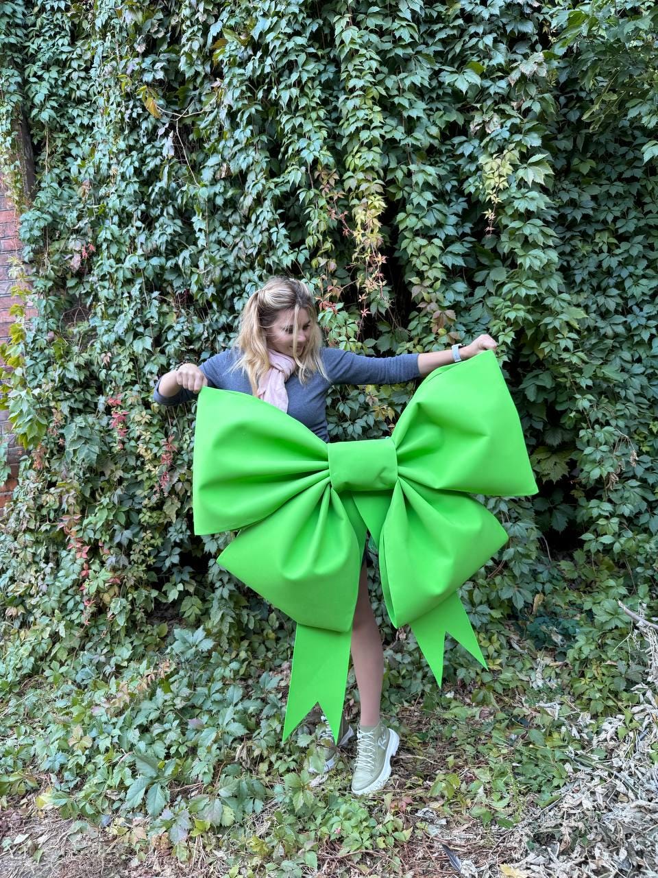 May include: A woman is holding a large, bright green bow in front of a wall of green ivy.