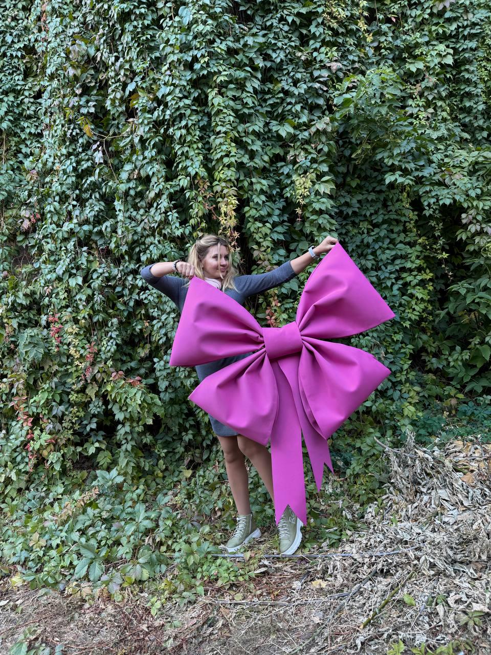May include: A large, bright pink bow made of fabric. The bow is being held by a person in front of a green leafy wall.