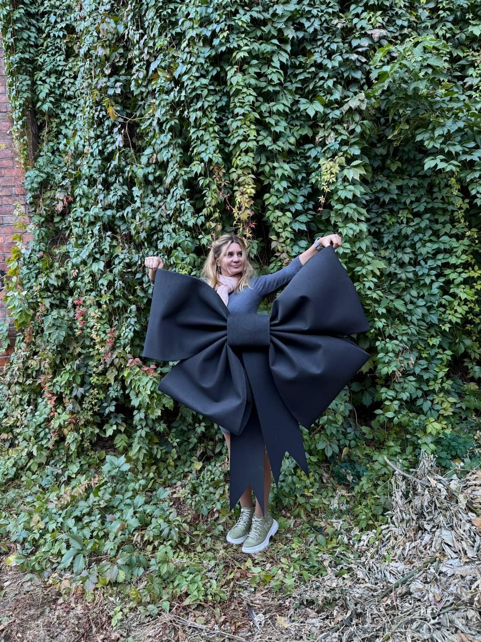 May include: A large black bow made of fabric, held by a person in front of a green ivy wall.