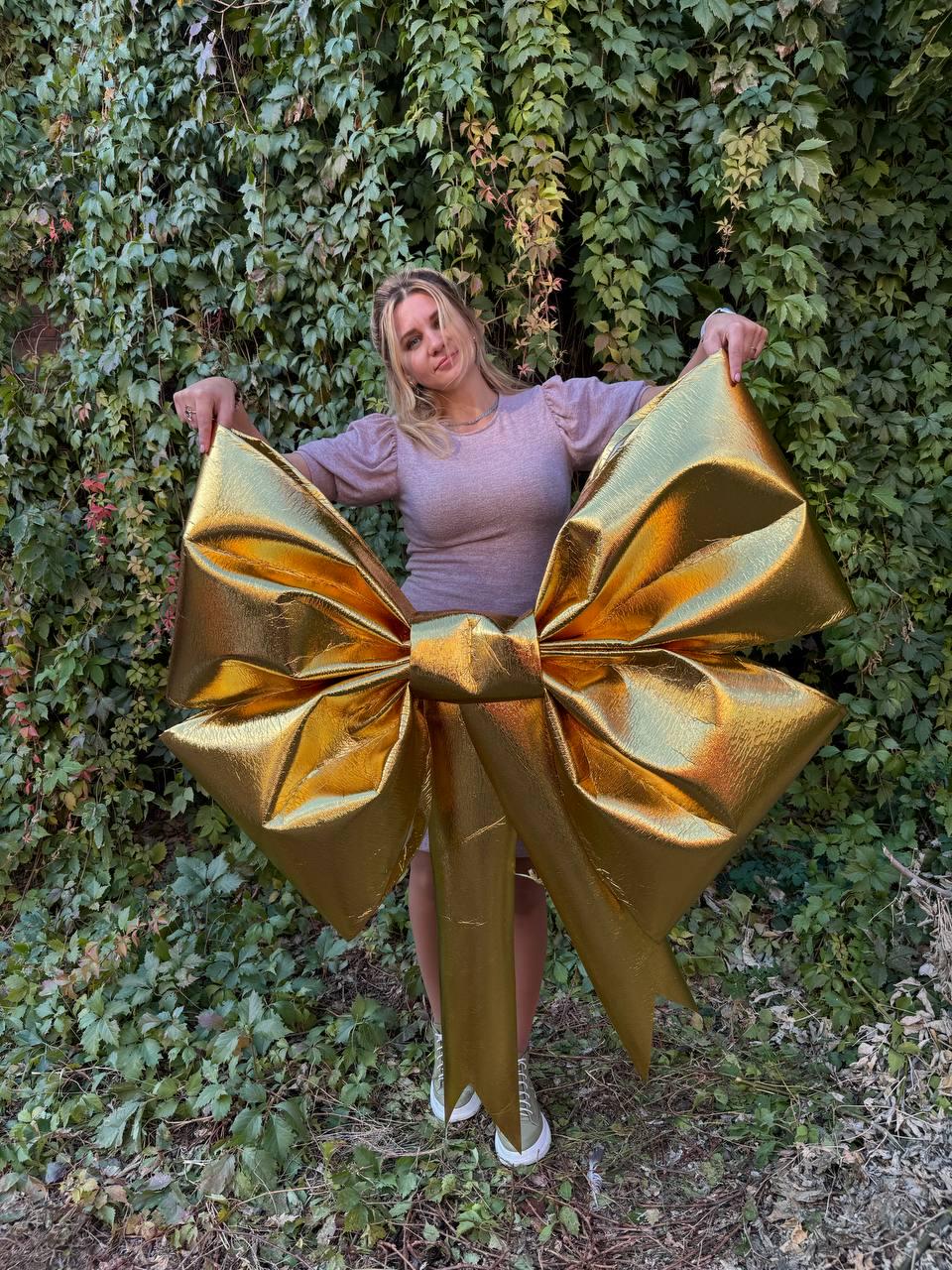 May include: A large gold bow made of fabric with a metallic sheen. The bow is being held by a person in front of a green leafy background.