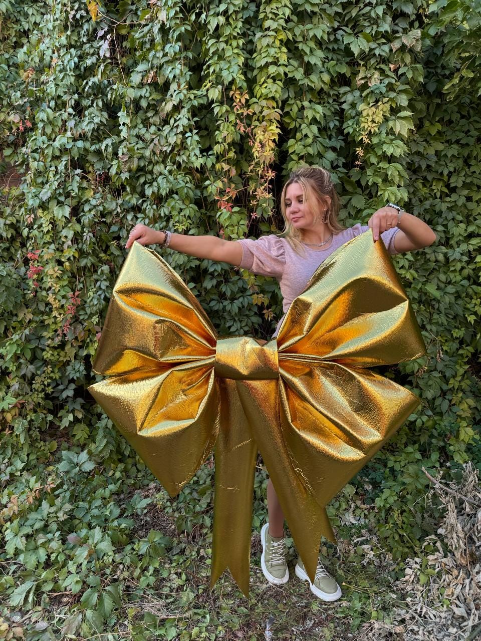 May include: A large gold bow with a metallic finish. The bow is being held by a person in front of a green leafy wall.