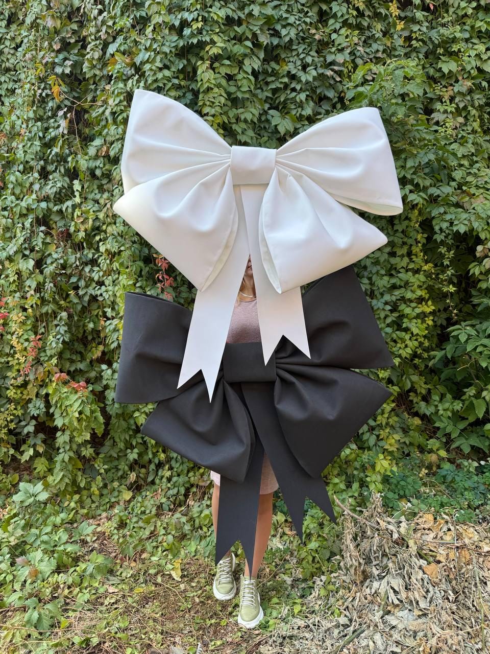 May include: A large white bow with a smaller black bow underneath. The bows are made of fabric and are tied together. The bows are in front of a green leafy background.