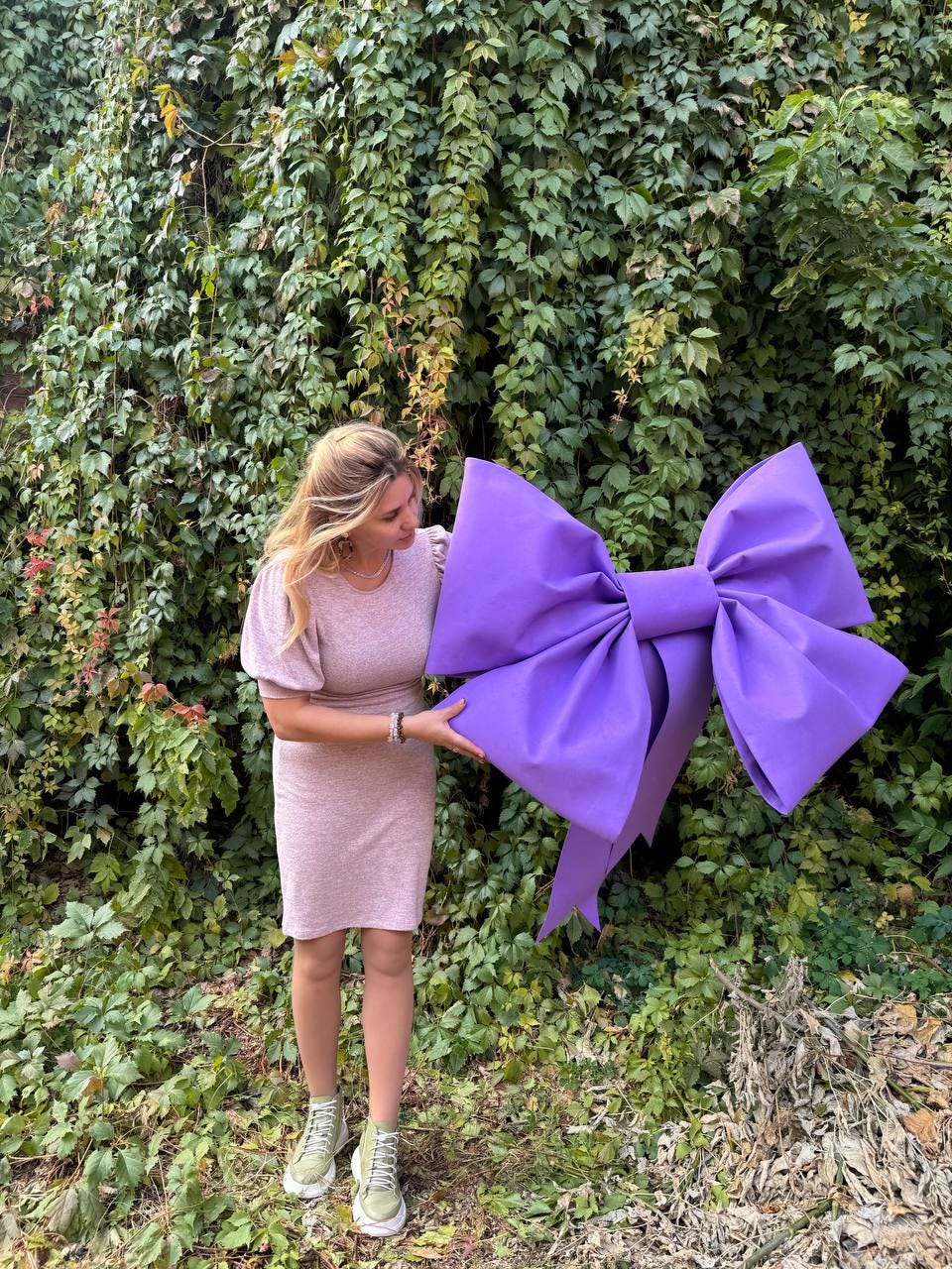 May include: A woman holds a large purple bow in front of a green leafy wall. The bow is made of fabric and has a large, rounded center.
