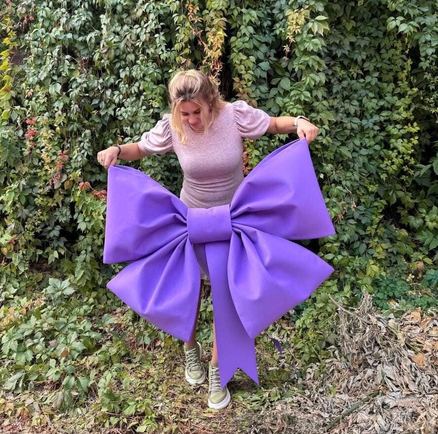 May include: A large purple bow made of fabric. The bow is so big that a person is standing in front of it, holding the ends of the bow.