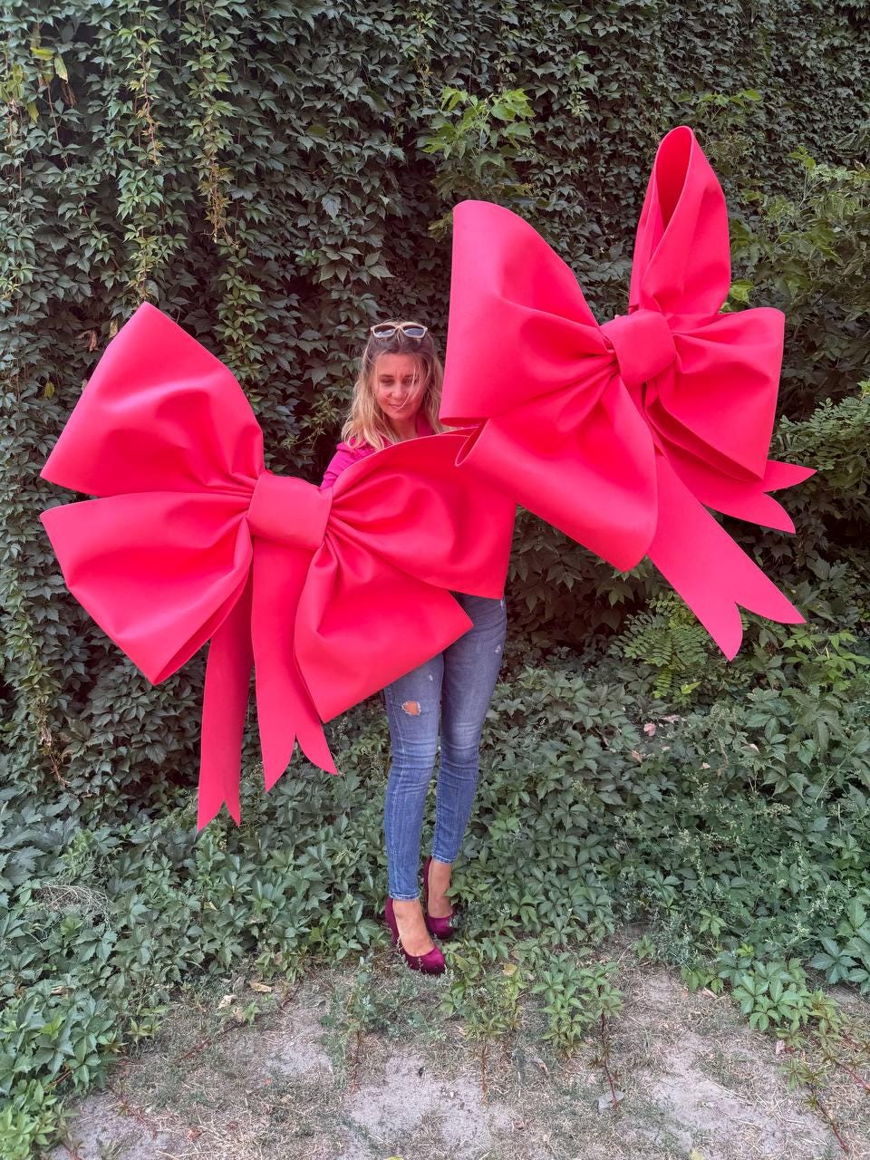 May include: Two large, bright red decorative bows. The bows are made of a smooth material and have long, flowing tails. The bows are in front of a person standing in front of a green leafy background.