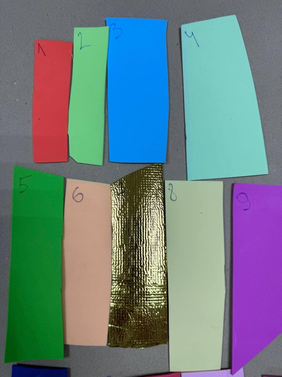 May include: A collection of nine rectangular paper swatches in various colors. The colors include red, green, blue, teal, green, peach, gold, light green, and purple. Each swatch is numbered with a black marker.