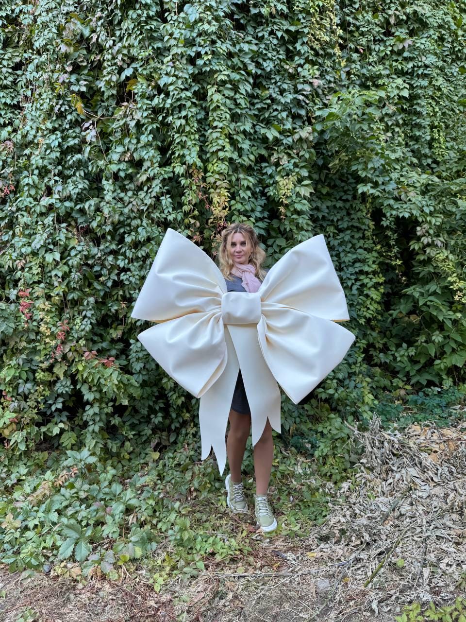 May include: A large white bow made of fabric, worn by a person standing in front of a green ivy wall.